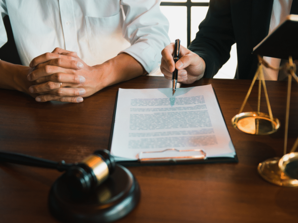 Two people sit at a desk with legal documents; one person is signing papers while the other folds their hands. A gavel and scales of justice are on the table.