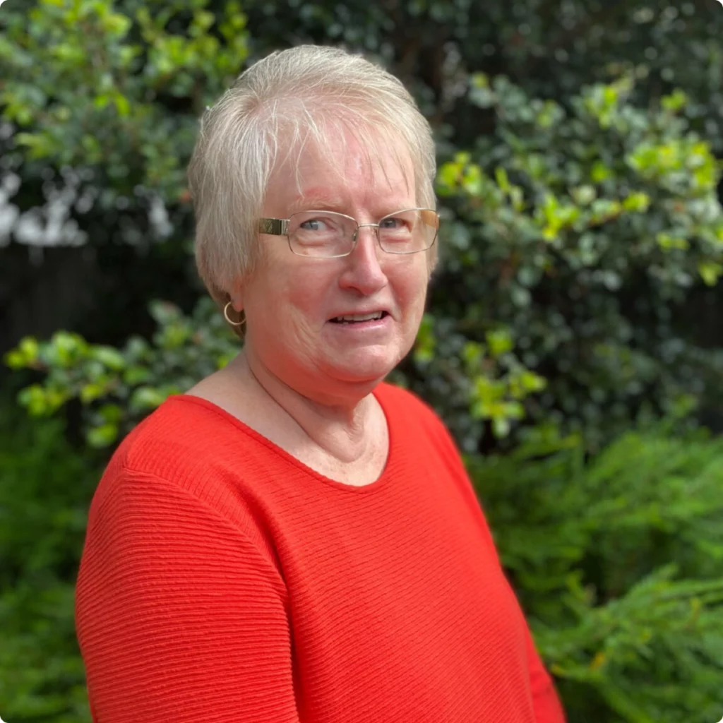 An older woman with short, light hair and glasses wears a red sweater and stands outdoors in front of green foliage.