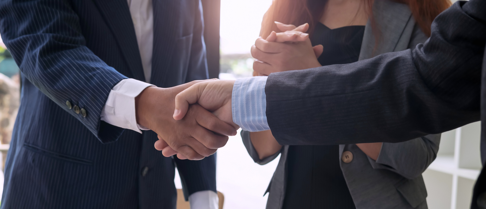 Two people in business attire shaking hands while a third person stands beside them with clasped hands, suggesting a business agreement or partnership.