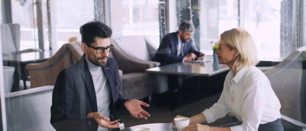 A man and a woman sit at a table in a cafe, having a conversation over coffee, while another man works on a laptop in the background.