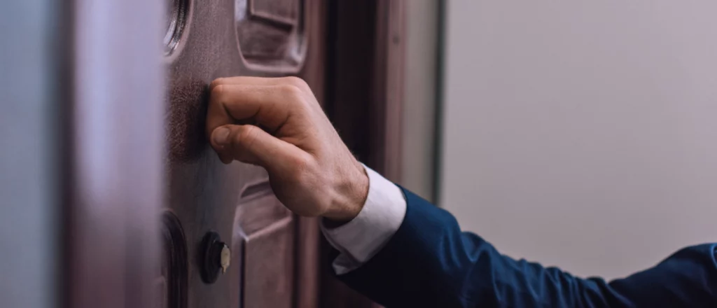 A person in a suit knocks on a wooden door with their right hand.